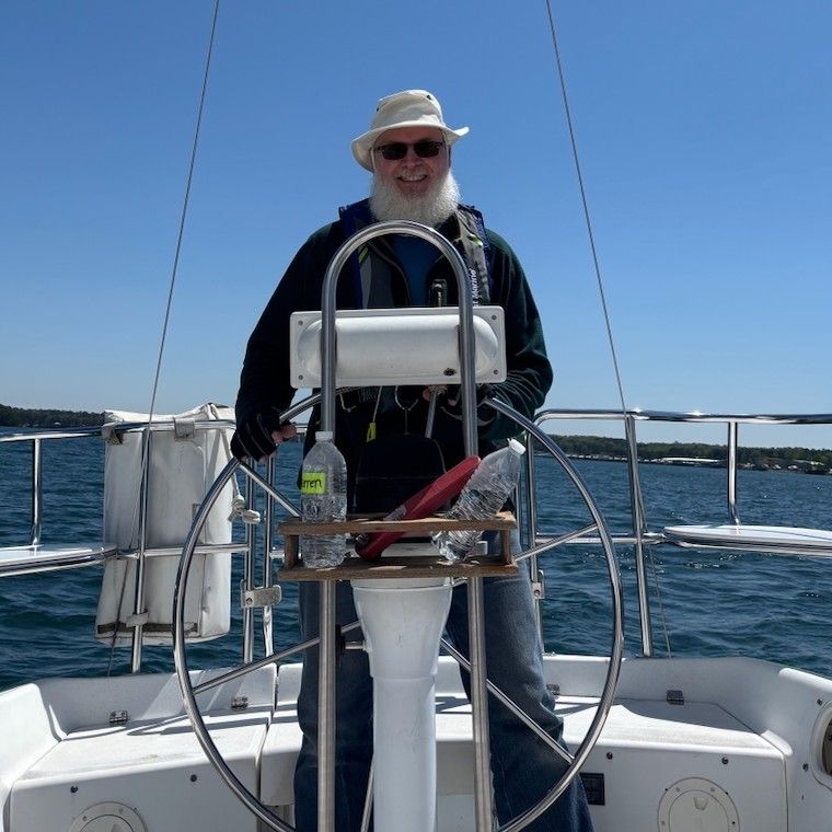 Darren at the wheel of a sailboat on open water, in a sun hat and sunglasses, smiling.
